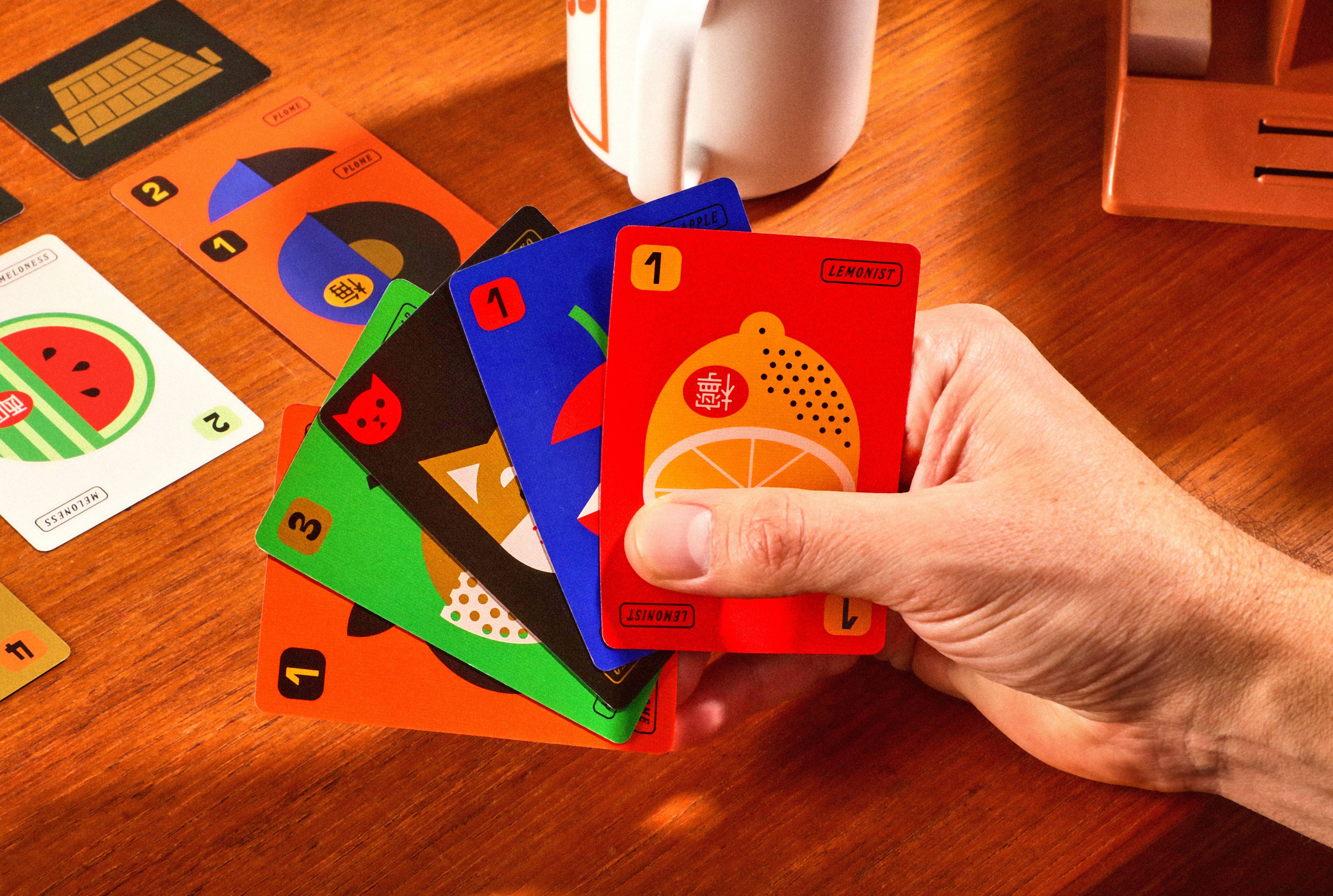 Fruit Boss cards held in a hand above a table, with additional cards and a mug blurred in the background.