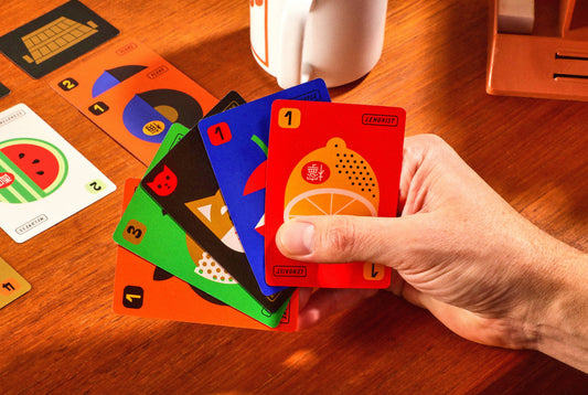 Fruit Boss cards held in a hand above a table, with additional cards and a mug blurred in the background.