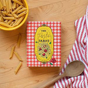 The Pasta Tarot Deck with penne, a wooden spoon, and a red-striped towel on a checkered cloth.