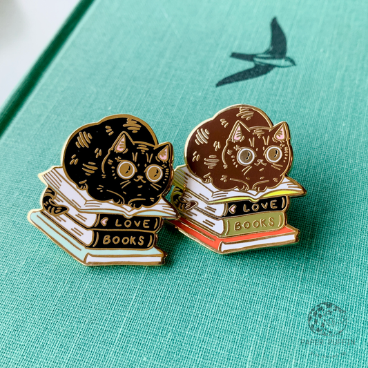 Two cat-shaped enamel pins, each resting on a stack of books labeled LOVE and BOOKS.