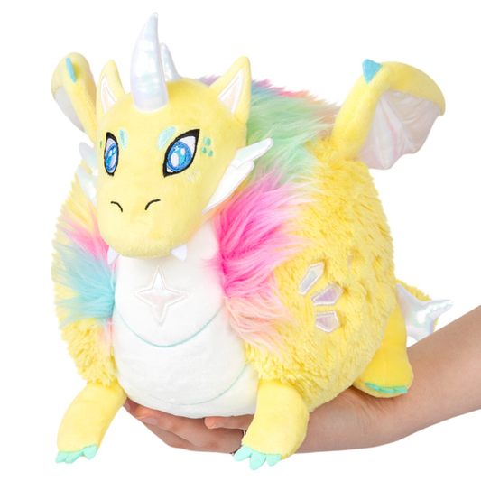 Mini Squishable Prismatic Dragon plush with rainbow fur, blue eyes, white horn, and wings held in hands.
