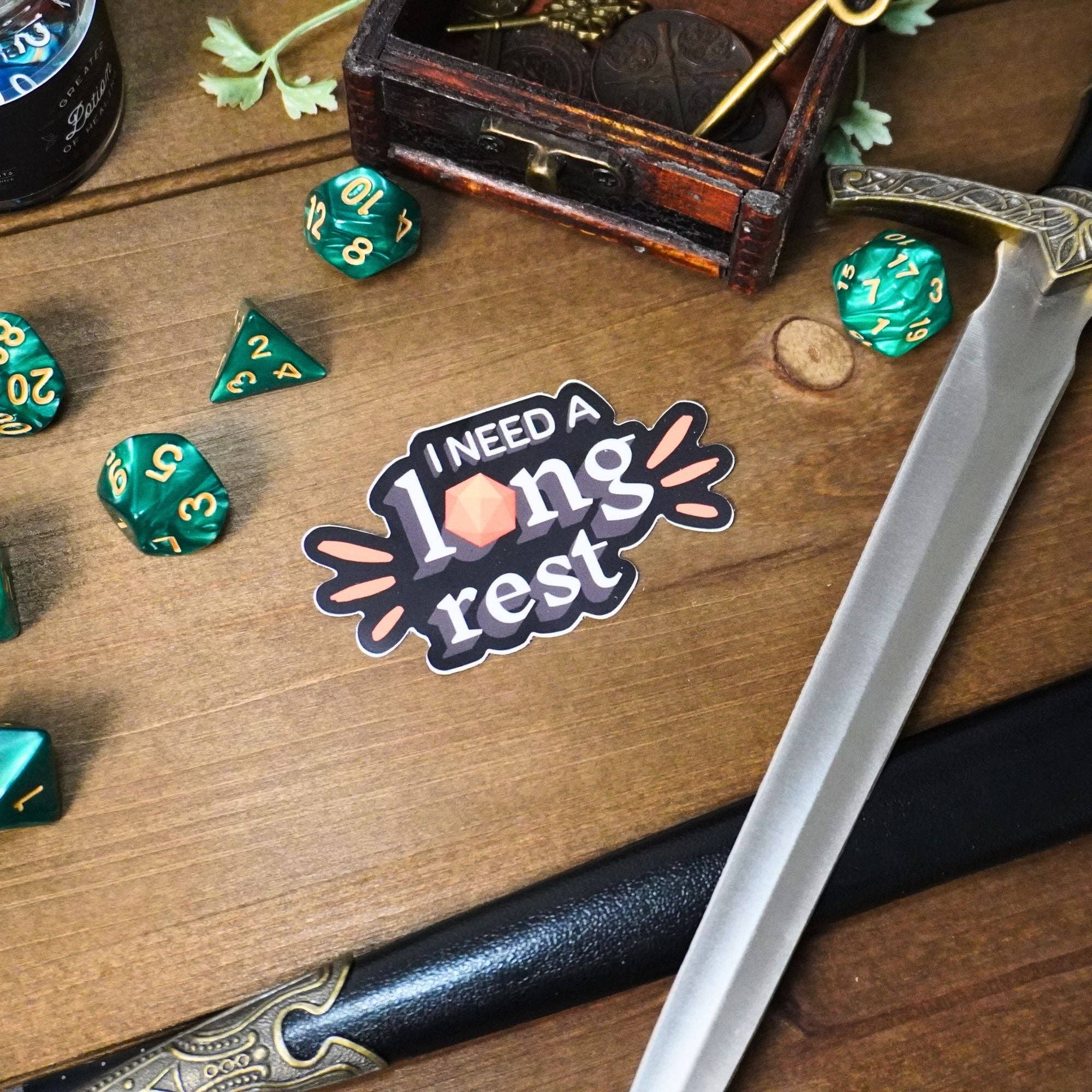 I Need a Long Rest D20 sticker shown with dice, sword, and small chest on wooden surface.