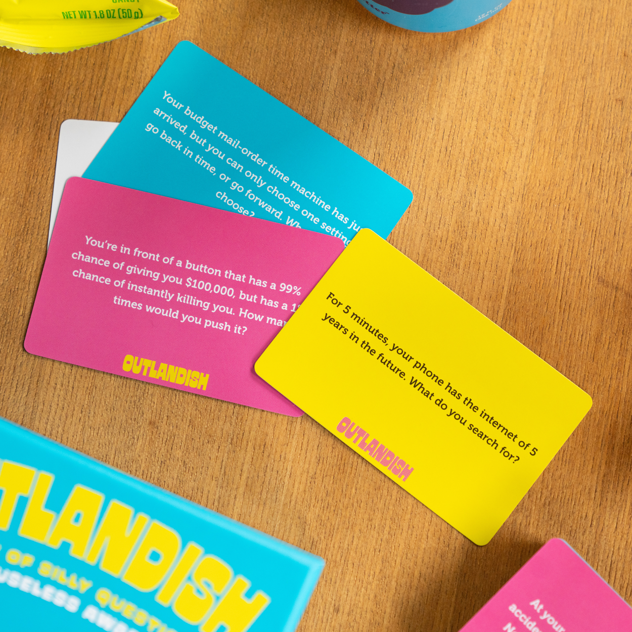 Three Outlandish game cards with colorful designs and question prompts displayed on a wooden table.