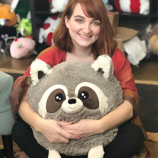 15 Raccoon II Squishable plush toy held by a woman with reddish hair sitting indoors.
