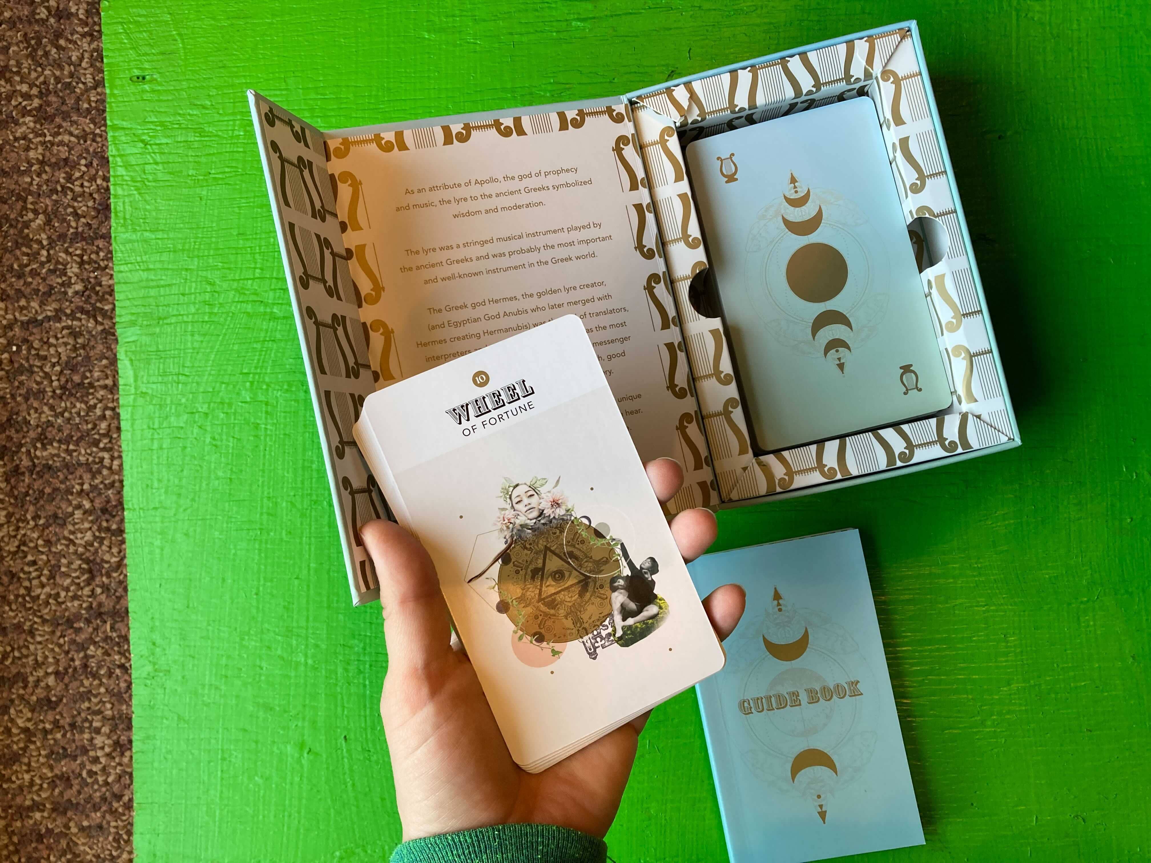 Gold Lyre Tarot Wheel of Fortune card held above its open box and guidebook on a green table.
