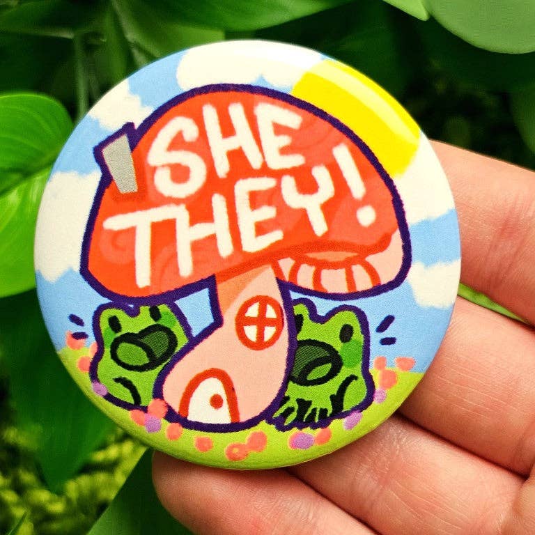 Large laminated waterproof button with SHE THEY text, two frogs, and a red mushroom design.