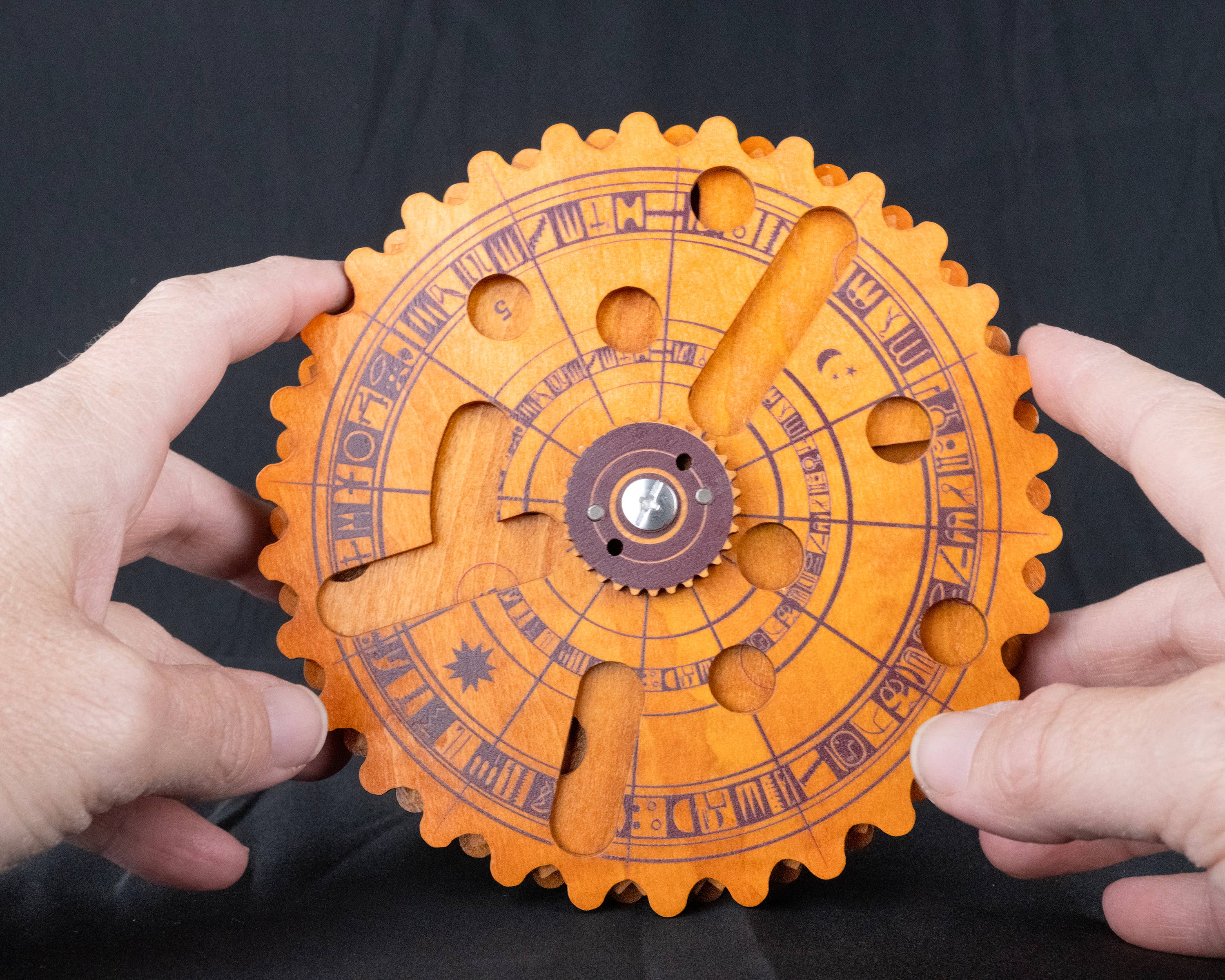 Egyptian Almanac Mathematical Brainteaser, round wooden puzzle with engraved symbols and gears, held by two hands.