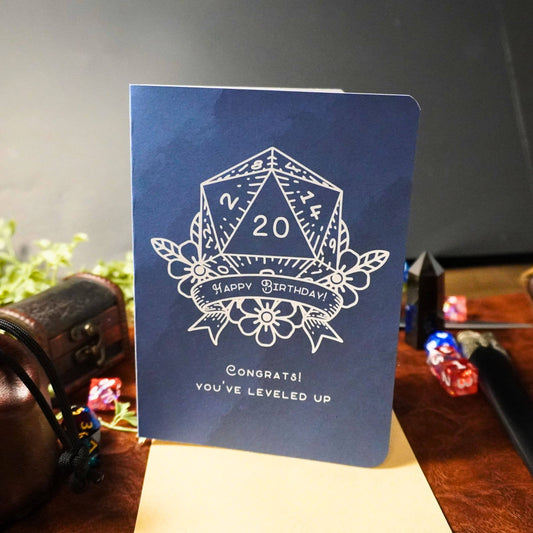 Blue D20-themed birthday card with white die, floral accents, and text: Congrats You’ve leveled up. Happy Birthday.