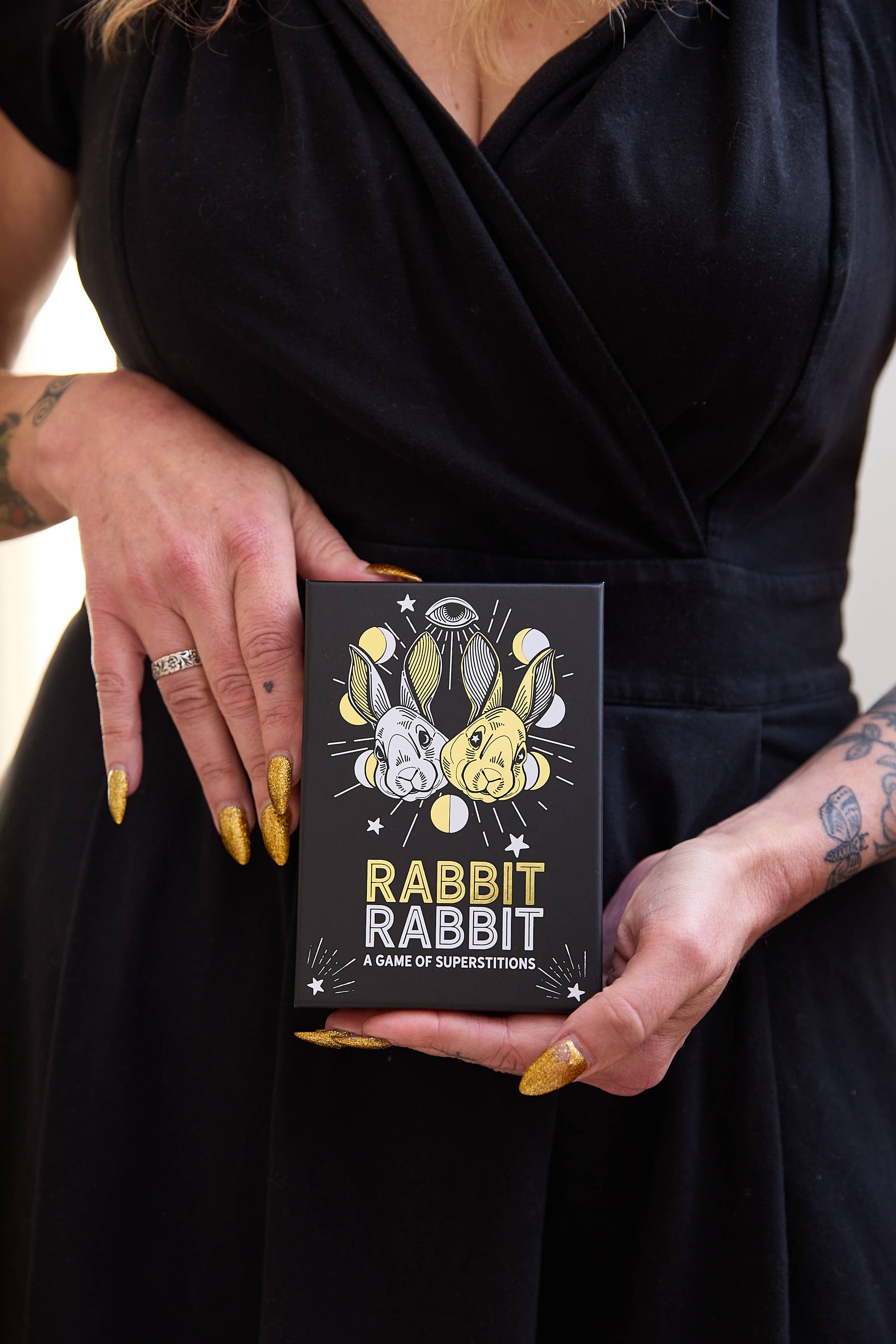 Rabbit Rabbit: A Game of Superstitions trivia game box held by a person with gold glitter nails.