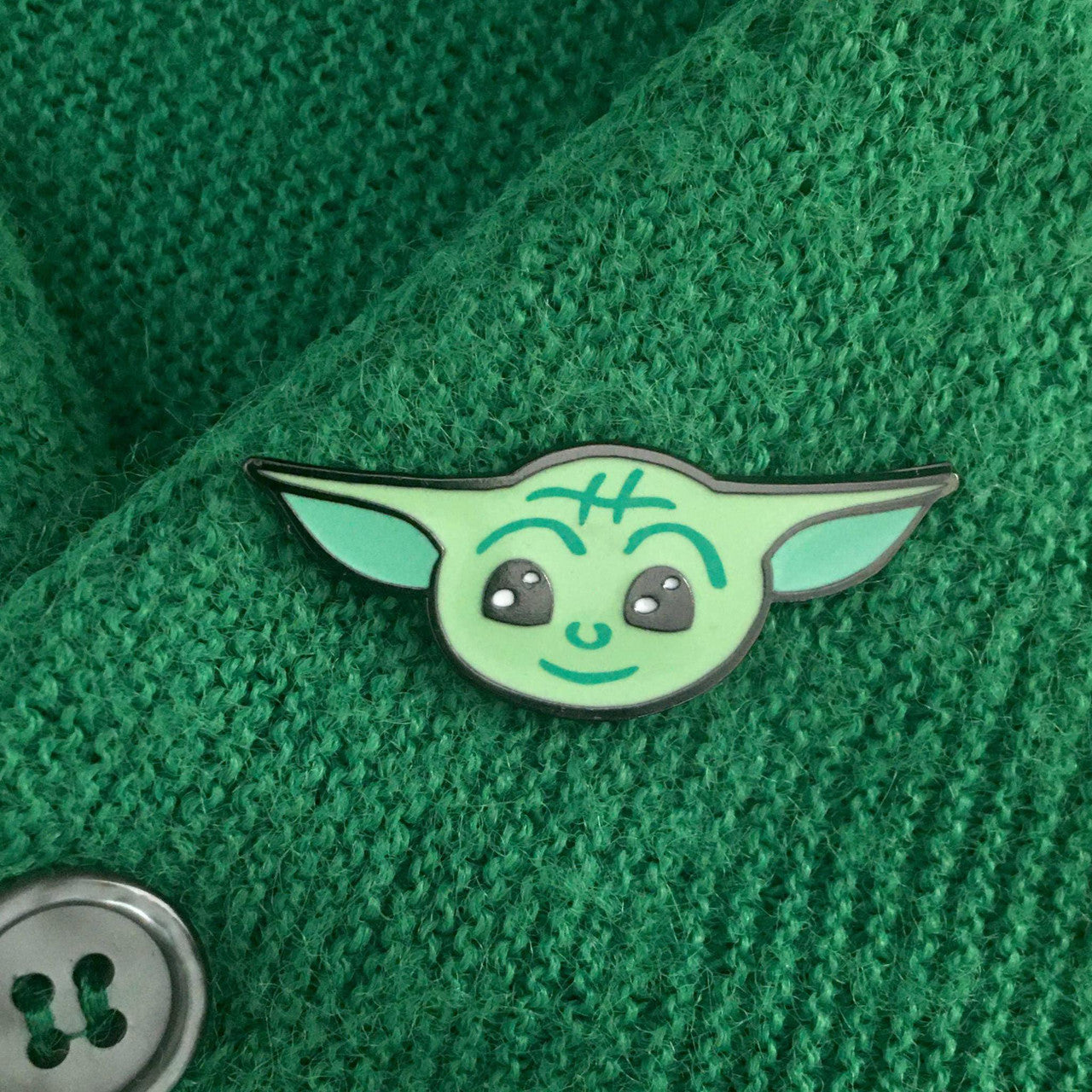 Baby Yoda enamel pin (Star Wars inspired) displayed on green fabric beside a large gray button.