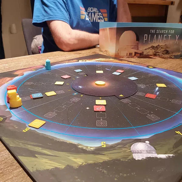 The Search for Planet X circular board game with colored tokens and a player seated at the table.