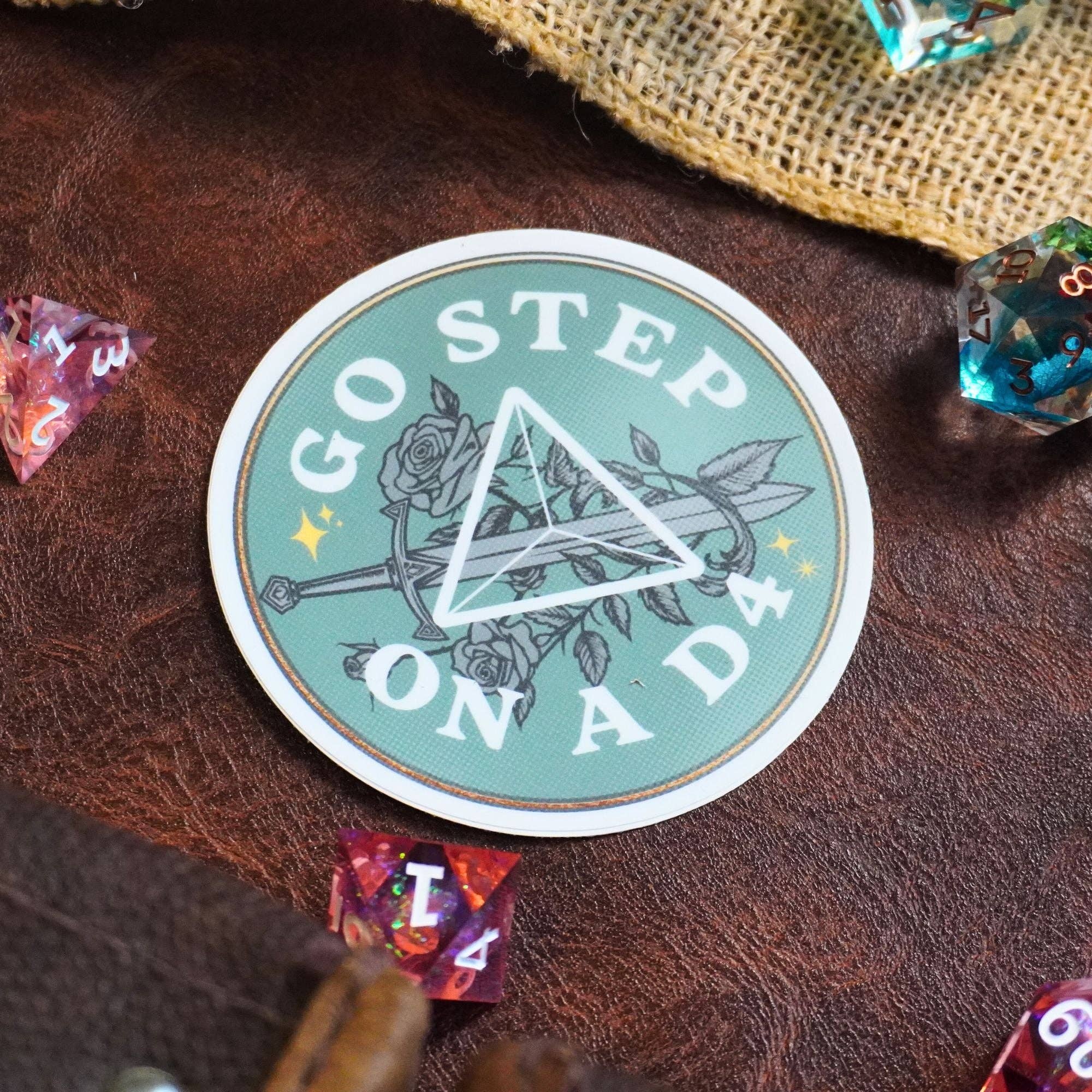 Go Step on a D4 Sticker featuring a floral D&D design with triangle shape, colorful dice, and leather-like details.
