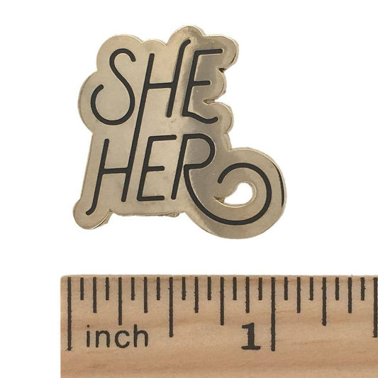 She/Her pronoun pin in black script beside a wooden ruler for scale.