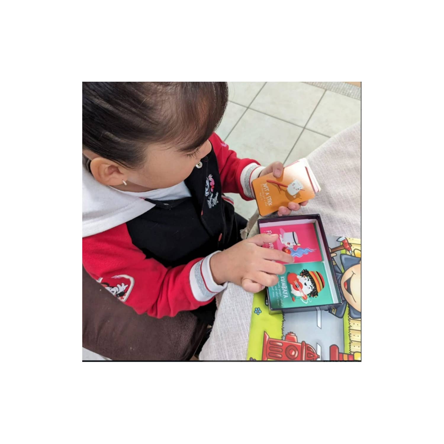 Child in red jacket playing Roast card game, with colorful cards on table.