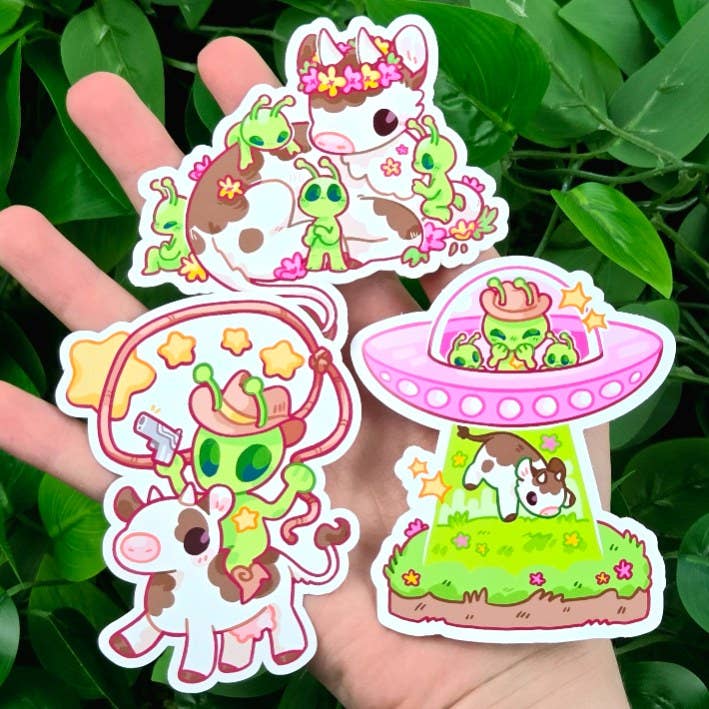 Cowboy Alien Stickers featuring four cows with space and farm themes, shown above green leafy plants.