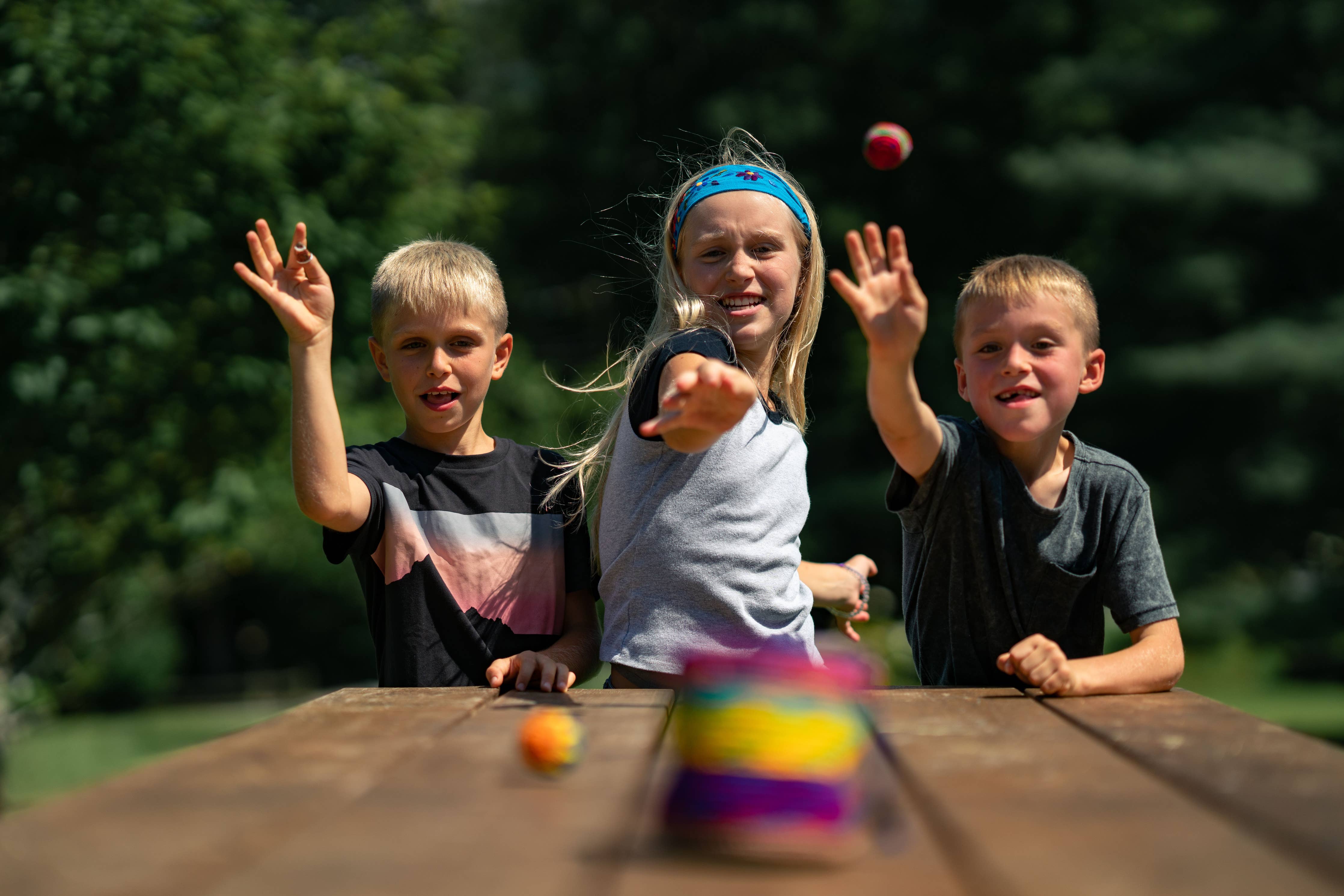 Canastaball Hacky Sacks being tossed by three children at stacked cans on a wooden table outdoors.