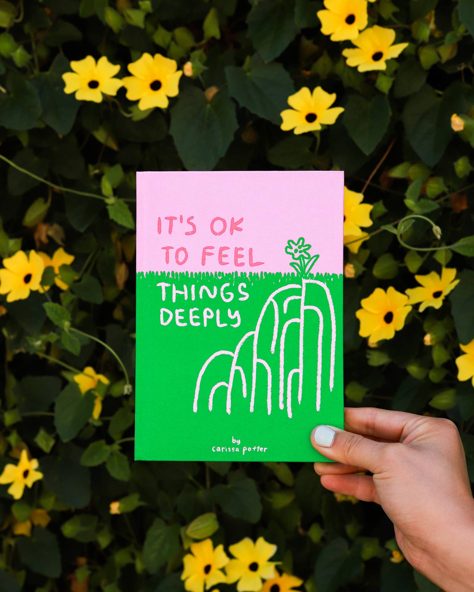 A hand holds the book It’s OK to Feel Things Deeply with yellow flowers and green leaves in the background.