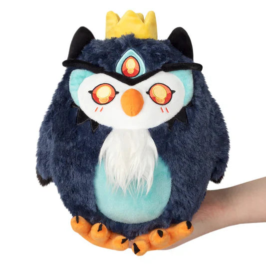 Mini Squishable Demon Owl plush toy, round and blue with a crown, orange feet, and glowing eyes, held in a hand.