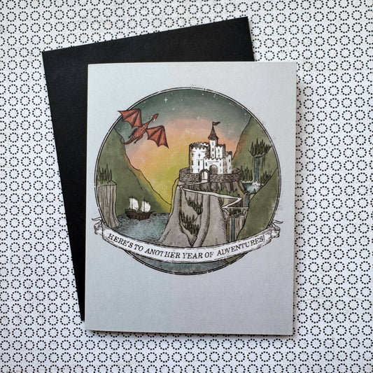 A5 fantasy birthday card with Heres to Another Year of Adventure text, castle, dragon, ships design; blank inside.