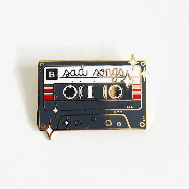 Sad Songs pin: a gold-plated enamel pin shaped as a black cassette tape with sad songs text and gold star accents.