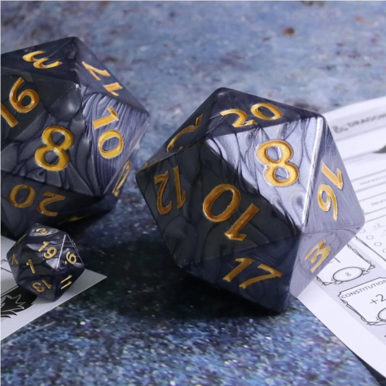Three 55mm Titan D20 Black Pearl dice with gold numbers on a table next to tabletop RPG character sheets.