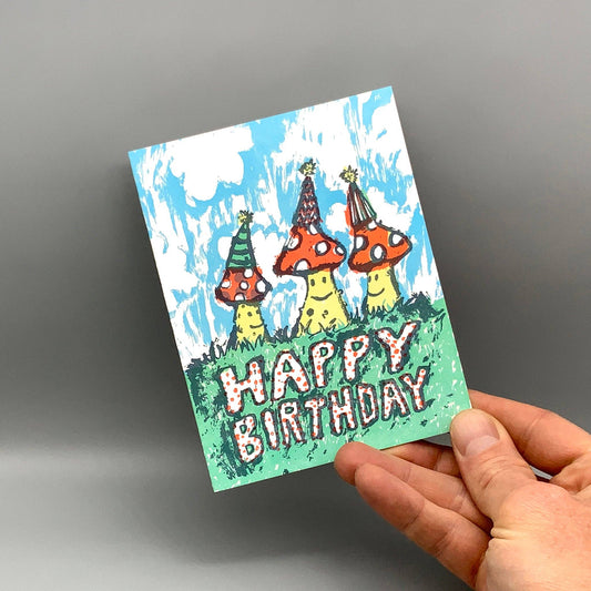 Birthday Mushrooms greeting card with three cartoon mushrooms wearing party hats on grass.