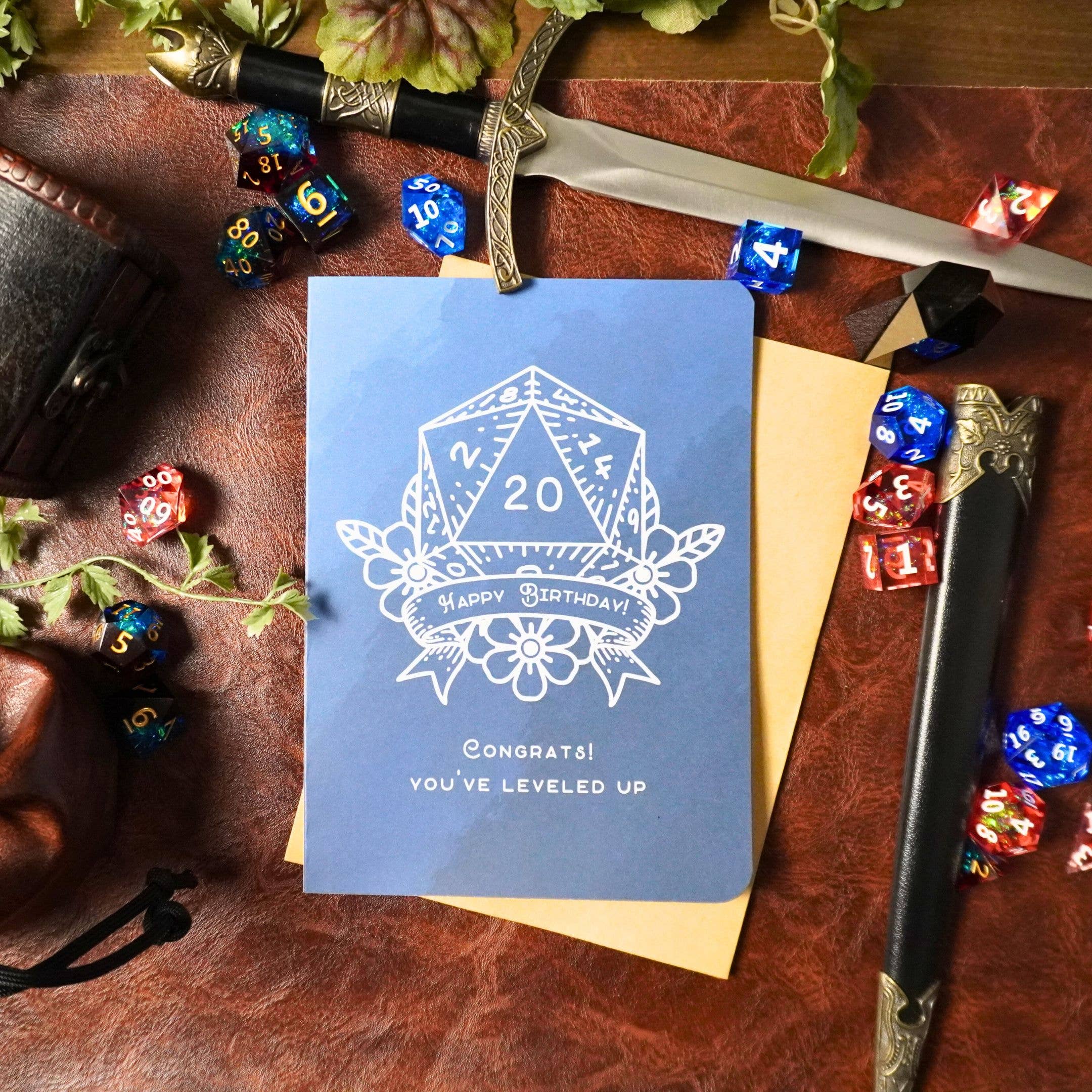 D20 Leveled Up DnD Birthday Card, blue with dice, sword, leather props. Text: Congrats You’ve leveled up.