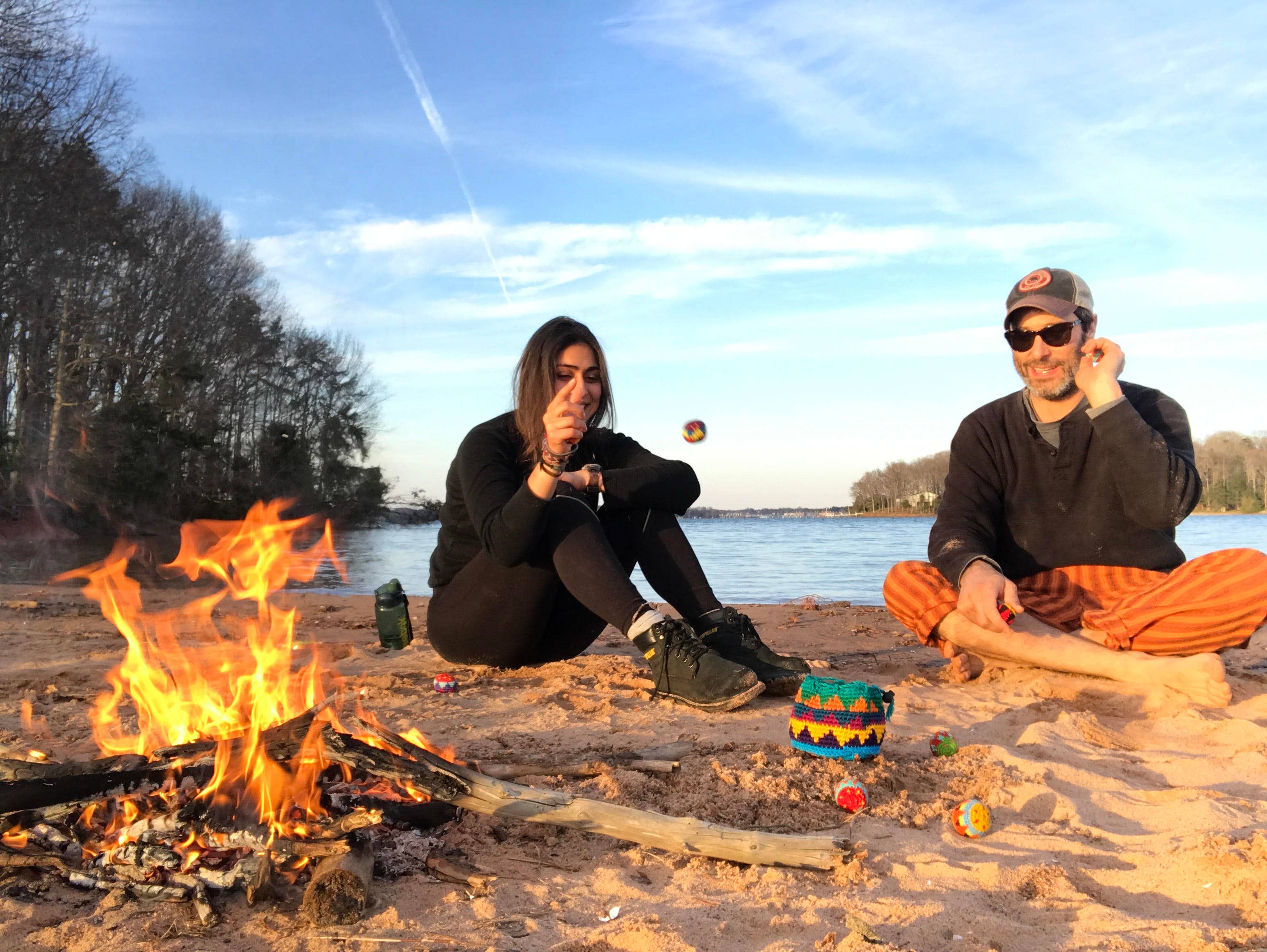 Two people use Canastaball Hacky Sacks by a campfire on a beach, with water and trees in the background.