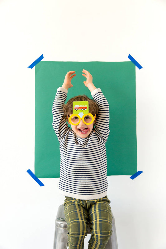 Child wearing yellow glasses for the What Am I Game, shown against a green paper backdrop.