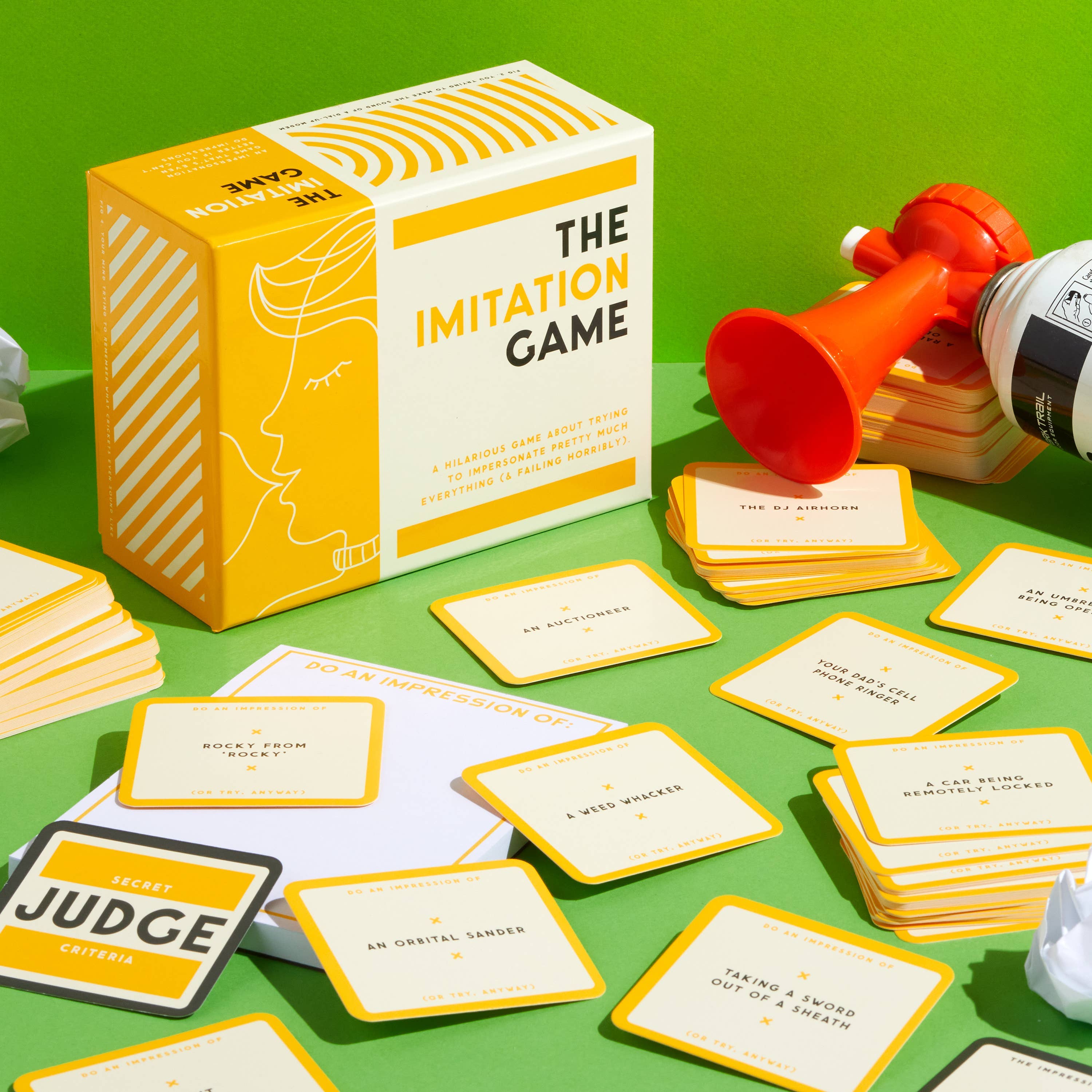 Brass Monkey The Imitation Game card set with judge pad, horn, and canister on green surface.
