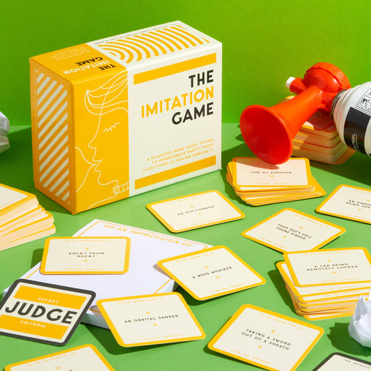 The Imitation Game party game with cards, red horn, and spray can displayed on a green background.