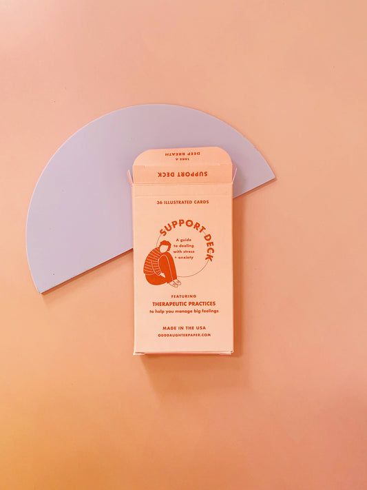 The Support Deck - Mental Health Card Deck in pink on a peach surface with a lavender semicircle backdrop.
