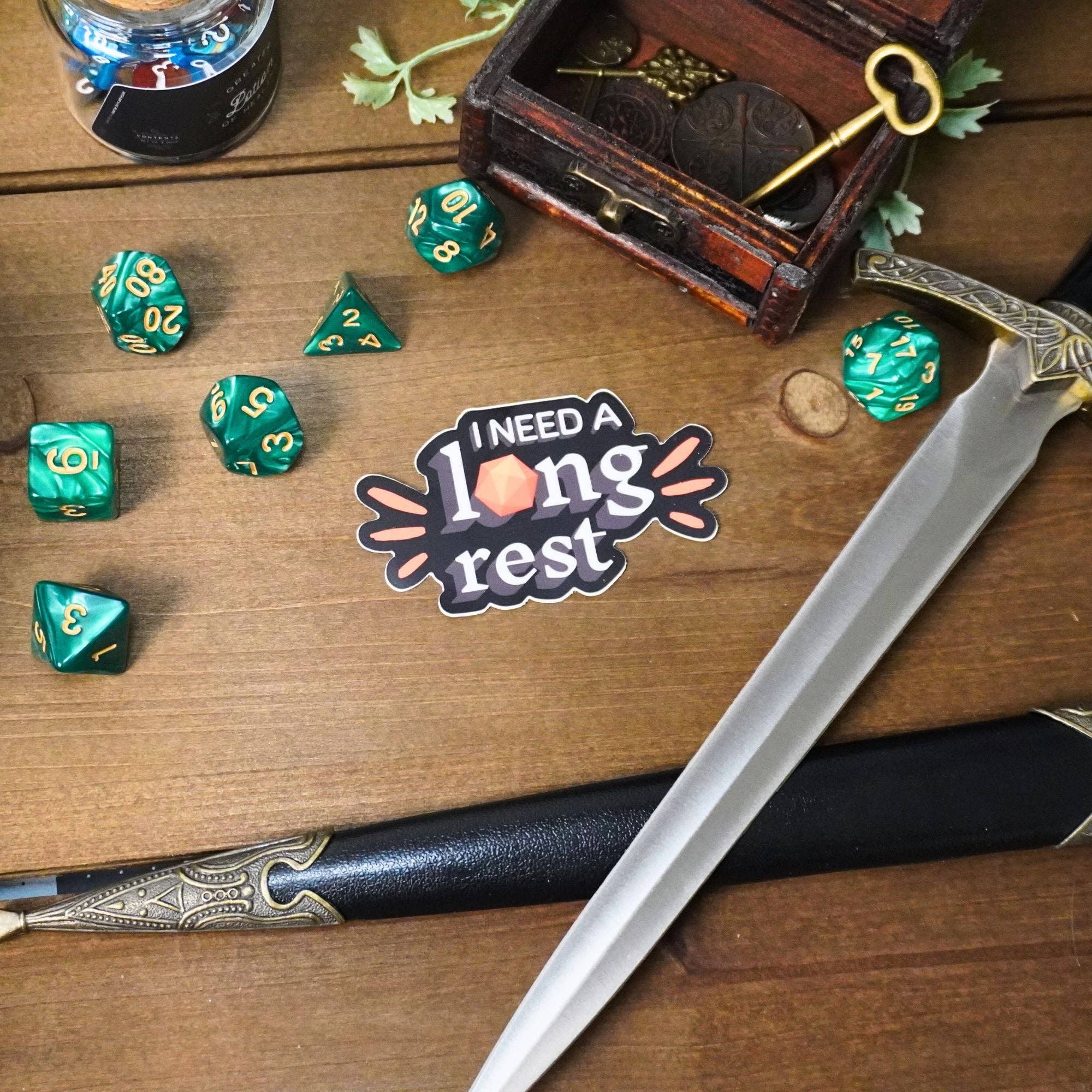 I Need a Long Rest Sticker with nearby green dice, a sword, and treasure on a wooden table.