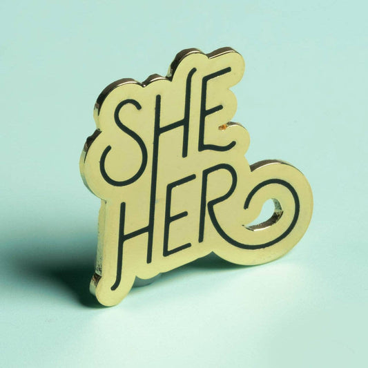 Gold She/Her pronoun pin with SHE HERO in black text on a light green background.
