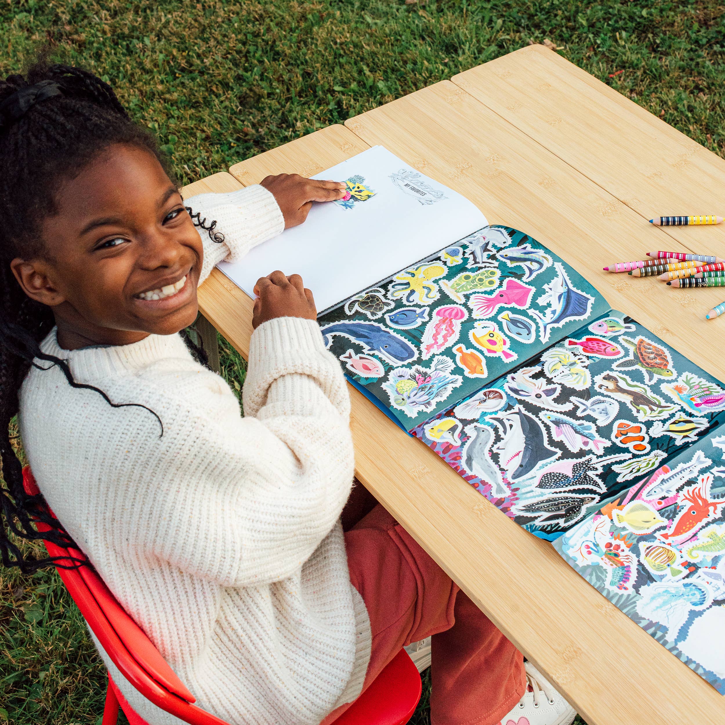 Child placing stickers in Learn to Draw Under the Sea with Stickers book at a wooden outdoor table.