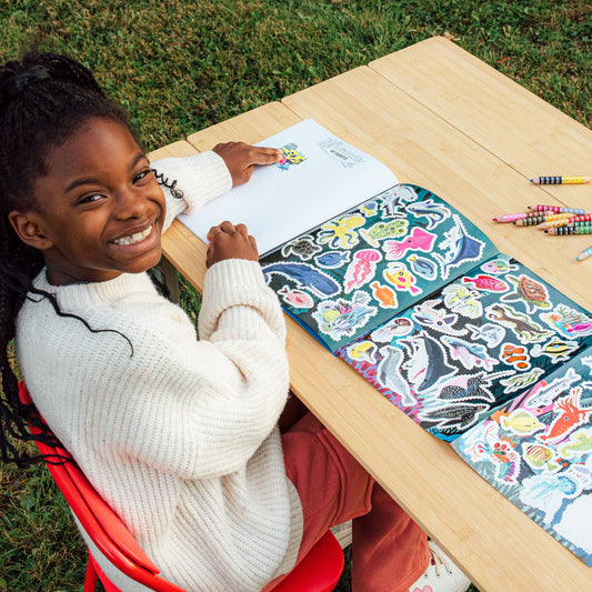 Child placing stickers in Learn to Draw Under the Sea with Stickers book at a wooden outdoor table.