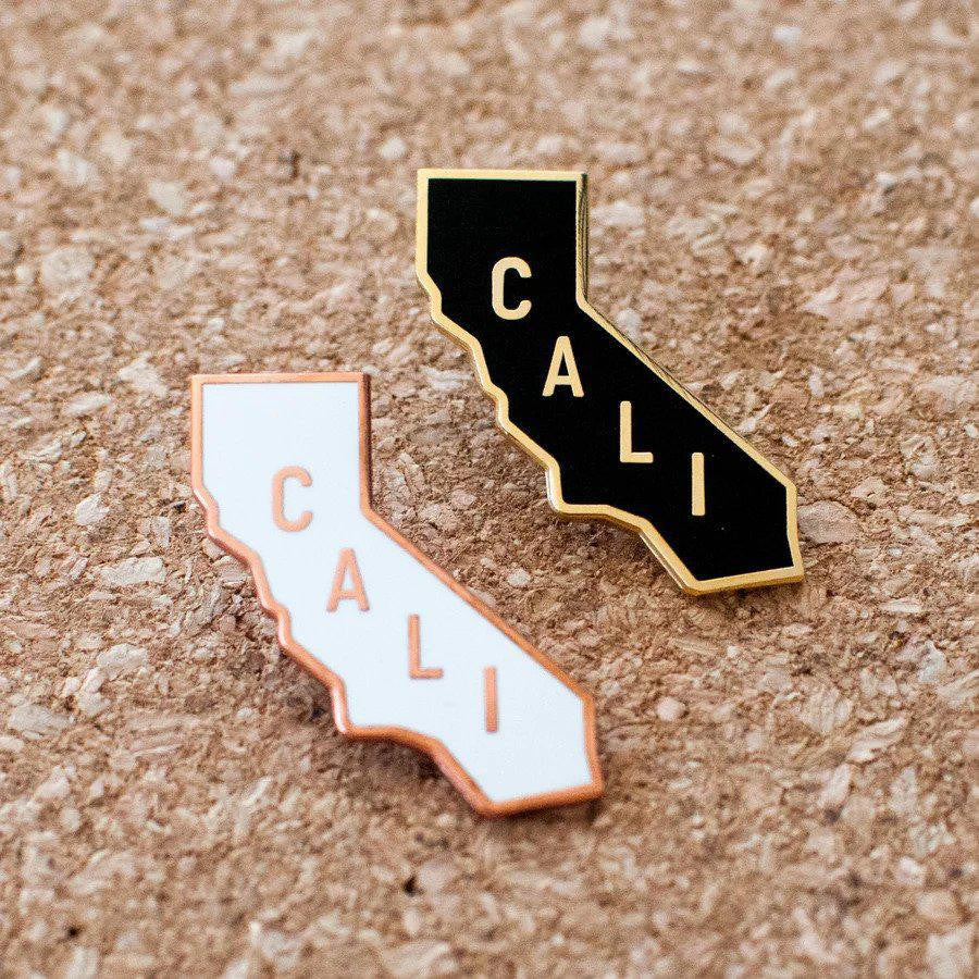 Two Cali Pins with CALI text, brass-plated edges, and rubber clutch backs displayed on cork.