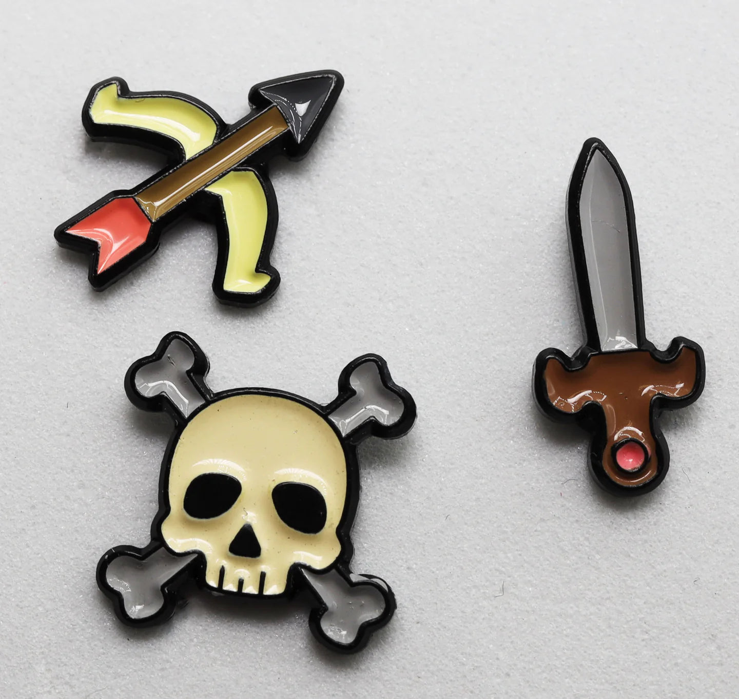 Set of three enamel pins featuring a bow and arrow, skull and crossbones, and a sword with distinct designs.