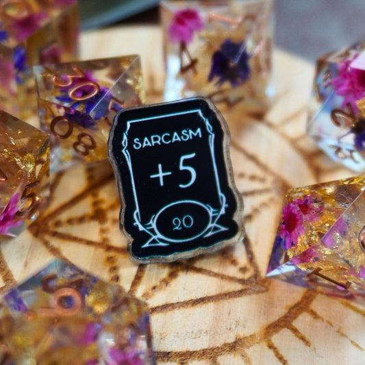 Sarcasm +5 Stat Acrylic Pin displayed with colorful resin dice on a wood-patterned surface.