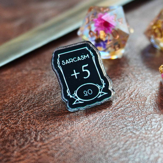 Sarcasm +5 Stat Acrylic Pin: book-shaped pin featuring dice and sword accents.