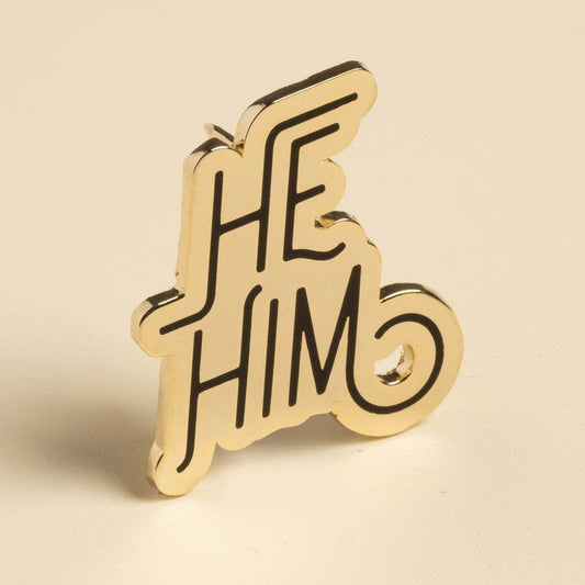 Gold enamel lapel pin with HE HIM in bold black text on a light beige background.