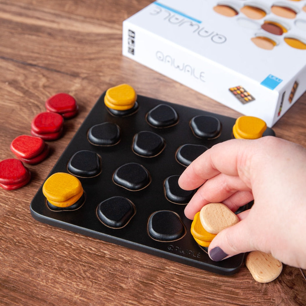 Qawale Mini wooden board with red, yellow, and black pieces; hand placing a piece. Box in background.