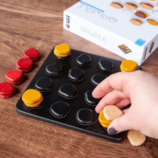 Qawale Mini wooden board with red, yellow, and black pieces; hand placing a piece. Box in background.