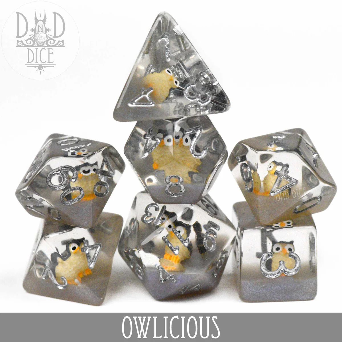 Gray resin 7-piece polyhedral dice set with small owl figures inside; Owlicious text visible. Dice stacked in a pyramid.