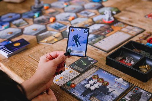 Scythe Expeditions Ironclad edition card held above a table with scattered game pieces and cards.