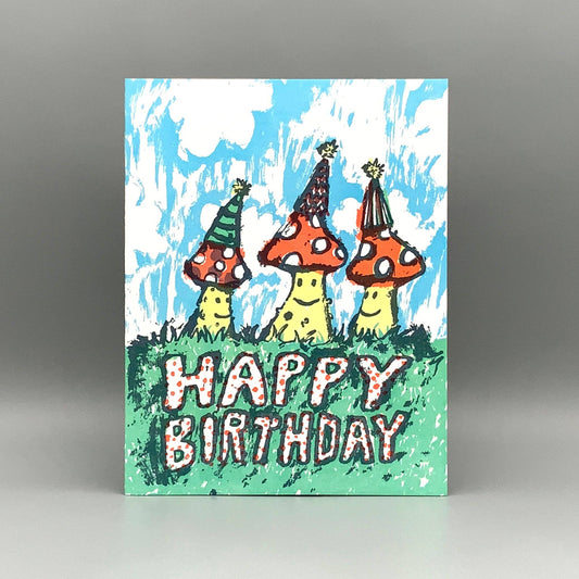 Birthday Mushrooms card featuring three cartoon mushrooms in party hats with HAPPY BIRTHDAY text on grass.