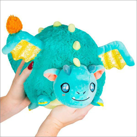 Mini Squishable Storybook Dragon plush (7), teal with yellow wings and orange-tipped tail, held by a person.