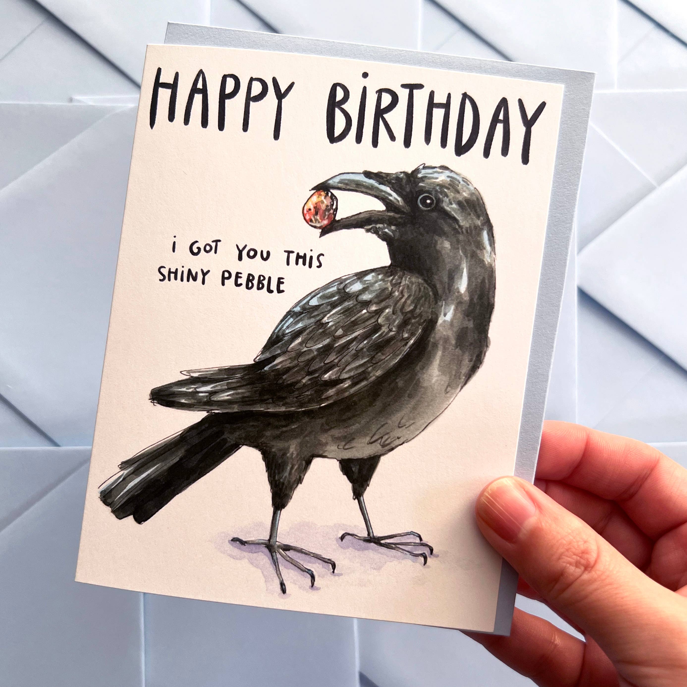 Crow I Got You This Shiny Pebble Happy Birthday Card held in a hand, showing a crow with a shiny pebble.