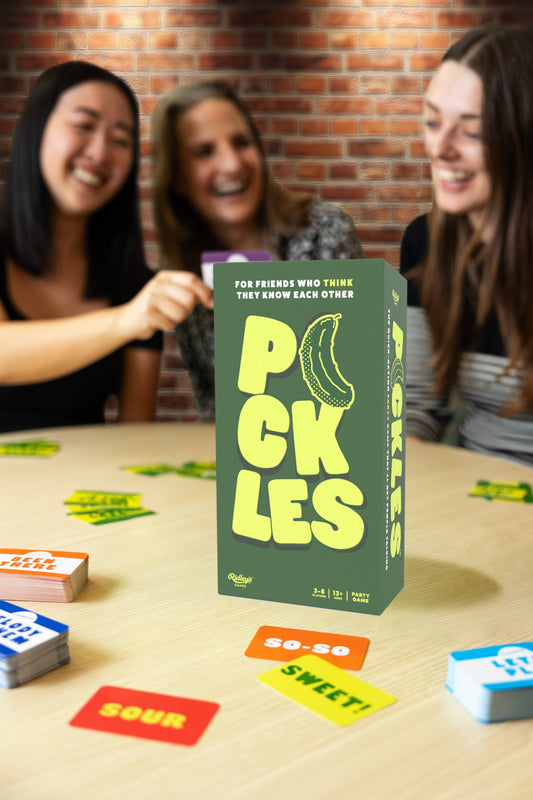 Three women play Pickles card game at a table with colorful cards and game boxes.