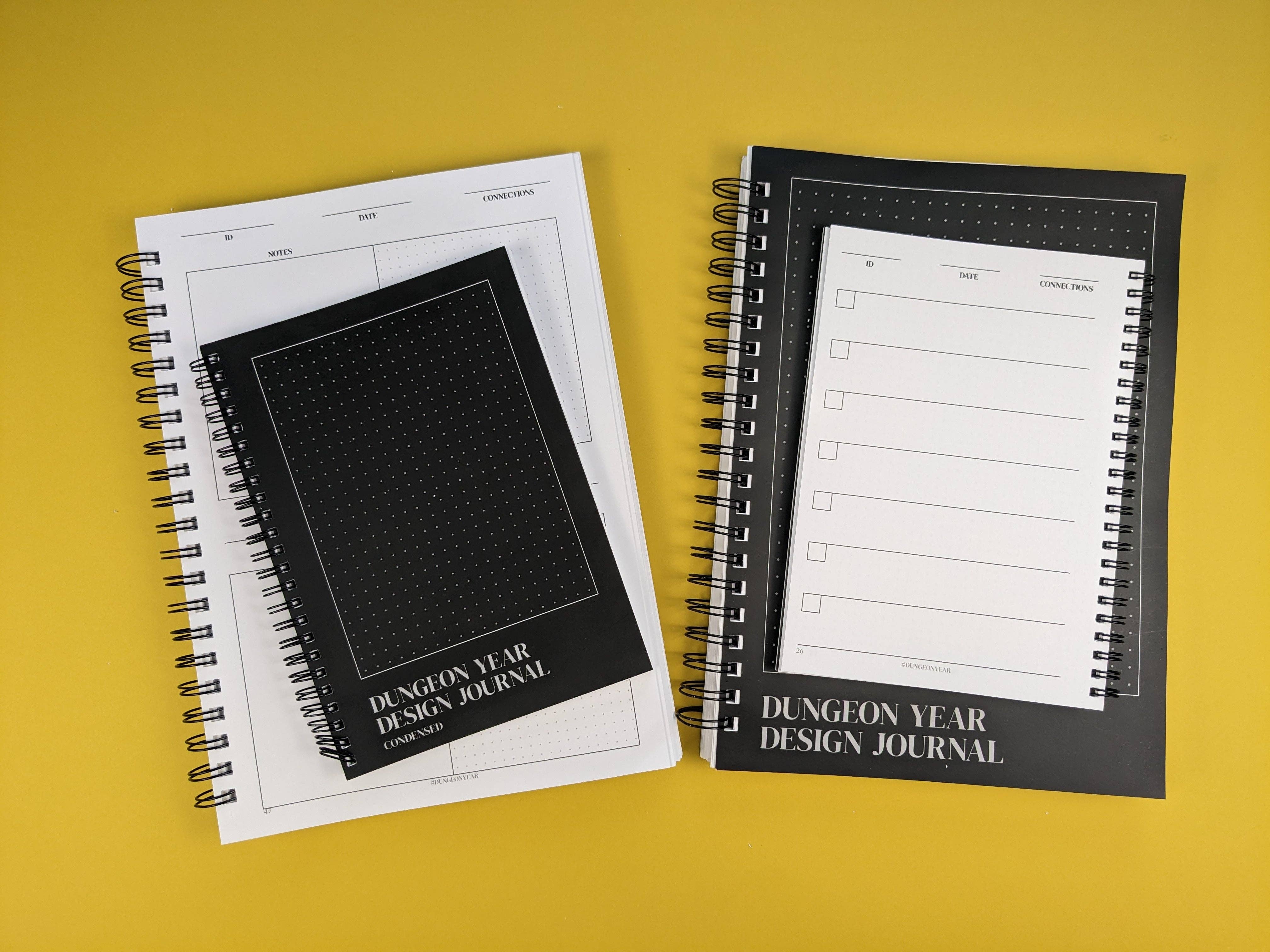 Large and small Dungeon Year Design Journals, both open and spiral-bound, on a yellow surface.